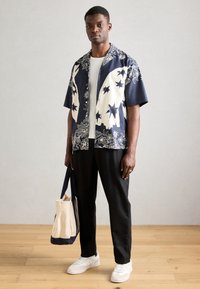 Κοντομάνικο ναυτικό μπλουζάκι με λευκά λουλούδια και σχέδια αστεριών, σε συνδυασμό με μαύρα παντελόνια και λευκά αθλητικά παπούτσια, κρατώντας μια μπεζ και ναυτική τσάντα.