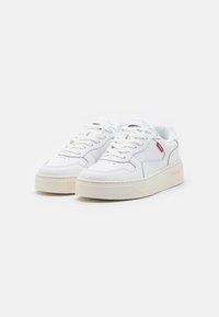 Zapatillas blancas con parte superior de cuero liso, punta redondeada, diseño de cordones, cuello acolchado y suela de goma en color crema contrastante. Logo de acento rojo.