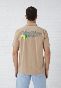Light brown short-sleeve shirt with a green and yellow lemon graphic and blue text on the back, featuring a straight hem and soft fabric.