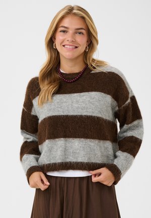 Femme souriante aux longs cheveux blonds porte un pull doux à rayures marron et grises, une jupe marron, un collier de perles violet et des boucles d’oreilles créoles.