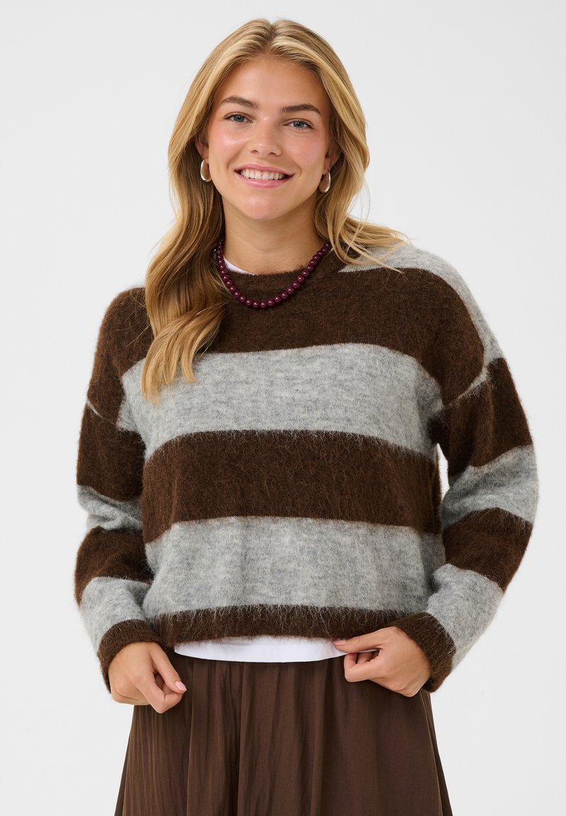 Femme souriante aux longs cheveux blonds porte un pull doux à rayures marron et grises, une jupe marron, un collier de perles violet et des boucles d’oreilles créoles.