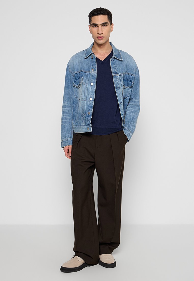 Giubbotto di denim azzurro chiaro, maglietta blu navy con scollo a V, pantaloni larghi marroni e scarpe beidge. Caratteristiche includono bottoni e dettagli delle tasche.