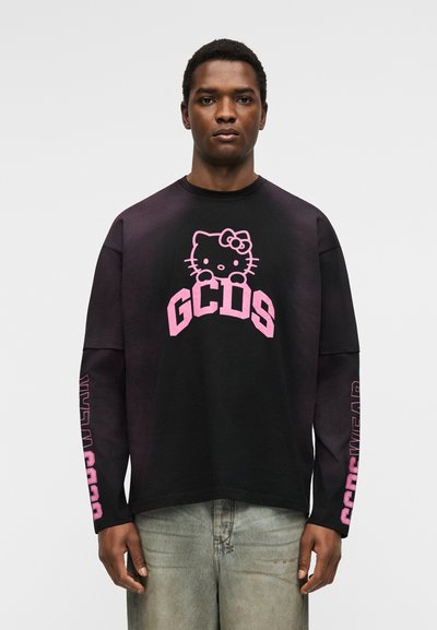 Άνδρας φορά μαύρο μακρυμάνικο πουκάμισο με ροζ γραφικό Hello Kitty και κείμενο "GCDS", συνδυασμένο με τζιν ανοιχτού χρώματος, στέκεται μπροστά από απλό φόντο.