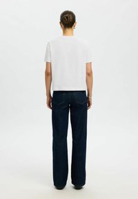 Femme aux cheveux en chignon portant un t-shirt blanc uni à manches courtes et un jean bleu foncé droit, debout de dos sur fond blanc.