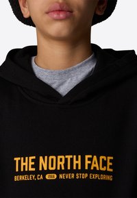 Felpa con cappuccio nera dal texture morbido, decorata con un logo giallo e il testo "THE NORTH FACE," Berkeley, CA, 1966, sul petto.