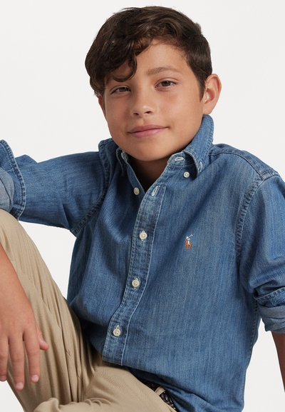 Τζιν πουκάμισο με κουμπιά σε ανοιχτό μπλε χρώμα, μακριά μανίκια, τσέπη στο στήθος και μικρή διακόσμηση με λογότυπο. Μπεζ παντελόνι ορατό μερικώς.