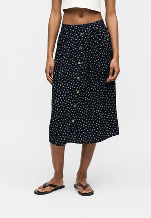 Mujer con falda midi negra con pequeños estampados florales blancos, botones frontales, top corto blanco y sandalias negras tipo flip-flop sobre fondo blanco.