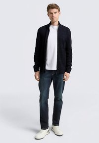 Marineblauwe gebreide cardigan met geribbelde structuur, ritssluiting, gedragen over een wit T-shirt, gecombineerd met donkere spijkerbroek en witte sneakers.