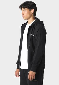 Felpa nera con zip e cappuccio, dotata di zip frontale e tasche laterali, indossata sopra una t-shirt bianca, esposta su uno sfondo grigio.