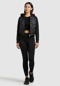 Kvinde iført sort cropped hættetrøje, sort vatteret vest med lynlås og hætte, sorte leggings og sorte sneakers, stående på en ensfarvet baggrund.