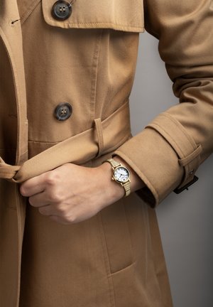 Manteau beige avec des détails de boutons, ceinturé, et une montre dorée avec un cadran blanc et un bracelet tissé, reposant sur une main.