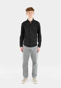 Svart langermet skjorte med krage som knyttes, parret med lysegrå denimbukser og beige sko, satt mot en ensfarget bakgrunn.