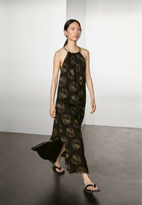 Sort maxikjole med halterneck, prydet med et stort cirkulært mønster i guld. Side-udskæring for let bevægelse. Båret med sorte sandaler.