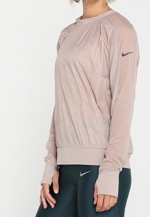 Vrouw die een licht mauve lange mouwen sporttop en donkere leggings draagt, beide met Nike-logo's, staande tegen een effen witte achtergrond.