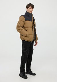 Giacca a vento marrone e nera, con colletto alto, cuciture orizzontali e tasche laterali. Indossata con pantaloni cargo neri e stivali.