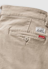 Bežna bombažna hlače z detajlom na zadnjem žepu, ki ima gumbasto zapiranje in rdečo oznako Levi's. Tkanina ima gladko, mat teksturo.