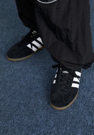 Des chaussures de sport en suède noir avec trois rayures blanches et des lacets noirs. Un pantalon noir texturé est partiellement visible à côté des chaussures.