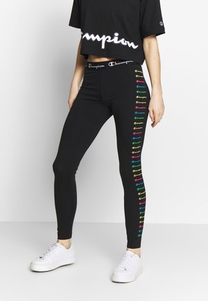 Person iført sort cropped Champion t-shirt og sorte leggings med farverige Champion-logoer ned ad det ene ben, kombineret med hvide sneakers.