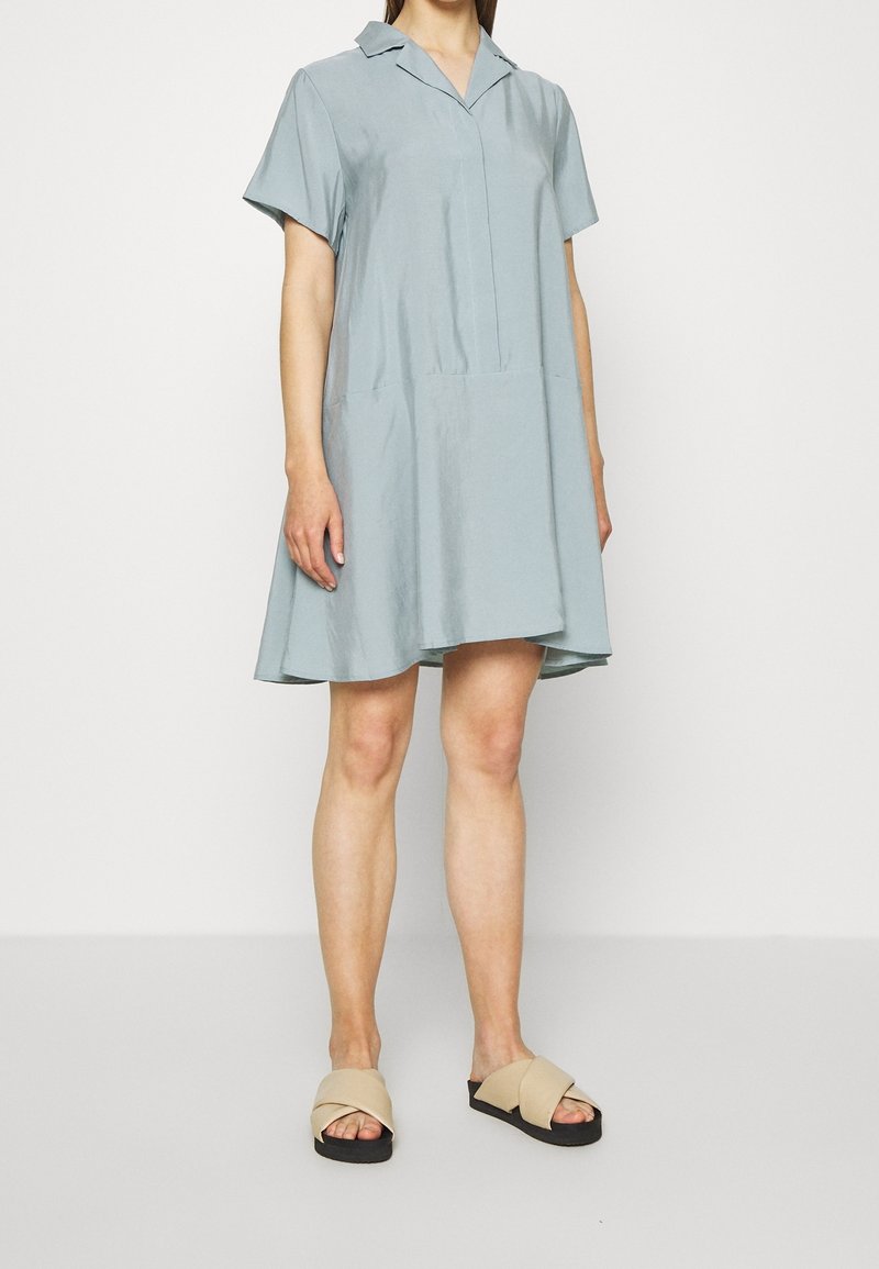 Robe ample à manches courtes en tissu bleu clair avec un col, présentant un design à panneaux et un ourlet évasé. Les sandales sont beiges avec une lanière croisée.