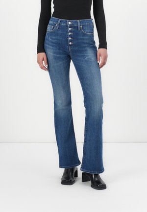 Augstas jostas zilu denimu flared džinsi ar pogu aizdari, pieciem kabatām un nedaudz izbalējuši. Apvienoti ar melniem zābakiem ar papēdi.