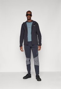 Sort jakke med hætte, lyseblå skjorte, marineblå bukser med grå detaljer og sorte sneakers. Glat stof og funktionelt design.