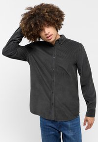 Camicia nera in corduroy con bottoni, maniche lunghe, colletto e orlo dritto, abbinata a jeans blu in denim. Dettaglio del tessuto testurizzato visibile.