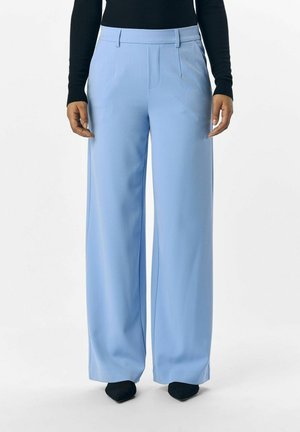Pantalons larges bleu clair avec des plis, fabriqués en tissu lisse. Présente une taille plate à l'avant et est assorti à des chaussures noires à bout pointu.