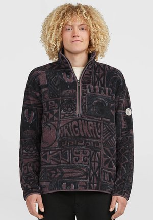 Person med krøllet blondt hår bærer en sort og lilla mønstret fleece med frontlynlås og brystlomme mod en ensfarvet baggrund.
