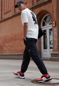 Mladý muž jde po chodníku, má na sobě bílý tričko s potiskem, černou čepici Nike, černé kalhoty a červeno-černé tenisky poblíž vstupu do cihlové budovy.