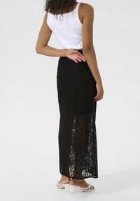 Gonna maxi nera in pizzo con un pannello trasparente, caratterizzata da motivi floreali e una vita aderente. Indossata con un top bianco attillato e sandali.