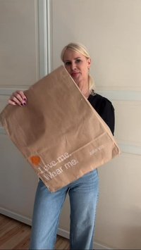 Ženska z blond lasmi drži veliko rjavo papirnato vrečko z napisom "Ljubite me. Nosite me." v svetli sobi z lesenim podom.