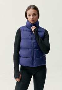 Mornarsko puhovko z visokim ovratnikom, z zadrgo spredaj in vodoravno prešito zasnovo; nosi se čez teksturirano črno majico z dolgimi rokavi in prilegajočimi se rokavi.