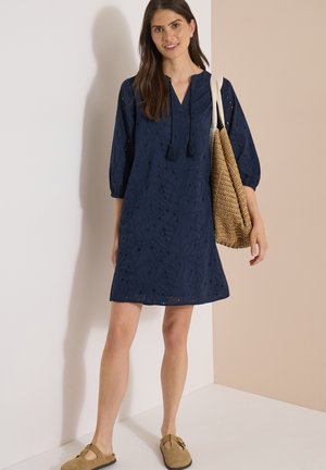Femme portant une robe bleu marine longueur genoux avec des manches trois-quarts, portant un sac fourre-tout tissé et des chaussures plates couleur beige.