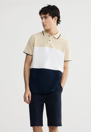 Polo a blocchi di colore beige, bianco e blu navy con colletto, patta con due bottoni e maniche corte. Indossato con pantaloni corti blu navy con texture.