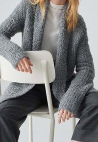 Persona con un cárdigan texturizado gris y pantalones oscuros se sienta hacia atrás en una silla blanca, con las manos descansando en el respaldo y en el costado de la silla.