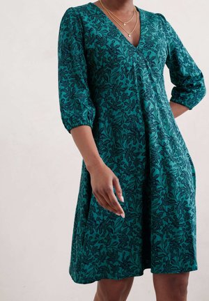 Femme portant une robe mi-longue bleu sarcelle avec un motif floral noir, des manches trois-quarts, et des colliers en or superposés avec des pendentifs croix.