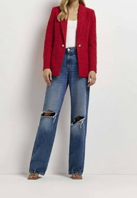 Femme portant un blazer rouge texturé, un haut blanc, un jean bleu déchiré et des sandales beige à bout ouvert sur un fond blanc uni.