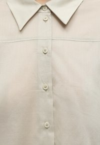 Gros plan d'une chemise beige à boutons avec un col pointu et quatre boutons visibles alignés verticalement.