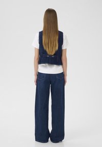 Sötét mosású farmerdzseki, melynek testhez álló fazonja van, hátul két gombbal, összeöltve széles szárú sötét farmernadrággal.