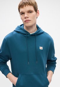 Žalias medvilnės mišinio su gobtuvu džemperis su kengūros kišene, veržlės su metaliniais galiukais ir siuvinėta logotipo etikete ant krūtinės.