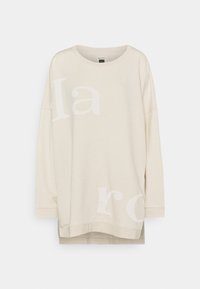 Felpa oversize color crema con maniche lunghe e scollo rotondo. Presenta una grande grafica testuale sbiadita sulla parte anteriore.