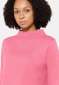 Femme aux cheveux bouclés naturels portant un pull en tricot rose à col montant, légèrement tournée vers la gauche sur un fond blanc uni.