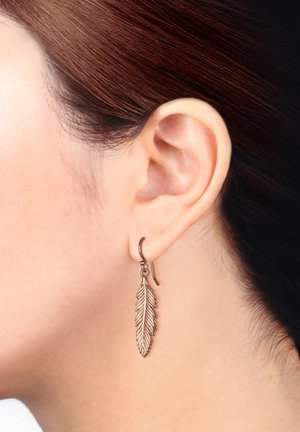 Boucles d'oreilles en bronze en forme de feuille, avec des détails fins et un design en crochet, suspendues juste en dessous du lobe de l'oreille. La surface texturée renforce l'intérêt visuel.