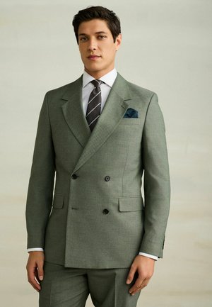 Mann trägt einen doppelt geknöpften grau-grünen Anzug, weißes Hemd, gestreifte schwarze Krawatte und dunknes Einstecktuch, steht vor einem einfarbigen Hintergrund.