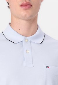 Gaiši zils polo krekls ar teksturētu audumu, aprīkots ar divu pogu aizdari, apkakli un mazu izšūtu logo uz krūšu kabatiņas.