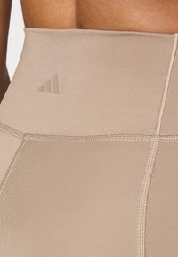 Beige høytliv leggings laget av stretchy stoff, med en teksturert overflate og subtilt tonet tredelt logo. Sømdetaljer synlige.