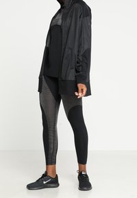 Completo sportivo nero composto da una felpa con zip, una maglia aderente e leggings con motivo e dettagli in rete, abbinato a scarpe da corsa nere.