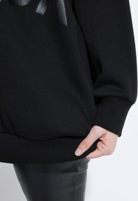 Zwarte sweatshirt met ribgebreide manchetten en onderkant, met een gestructureerd ontwerp, waarbij een hand de onderrand vasthoudt. Glad stofoppervlak.