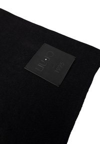 Tessuto di feltro nero con superficie testurizzata e un'etichetta in pelle nera rettangolare che presenta testo in rilievo e una decorazione a borchietta.