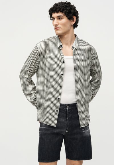 Νέος με κατσαρά μαλλιά φοράει ασπρόμαυρο πουκάμισο με καρώ πάνω από λευκό φανελάκι και μαύρα τζιν σορτς, με τα χέρια πίσω από την πλάτη.